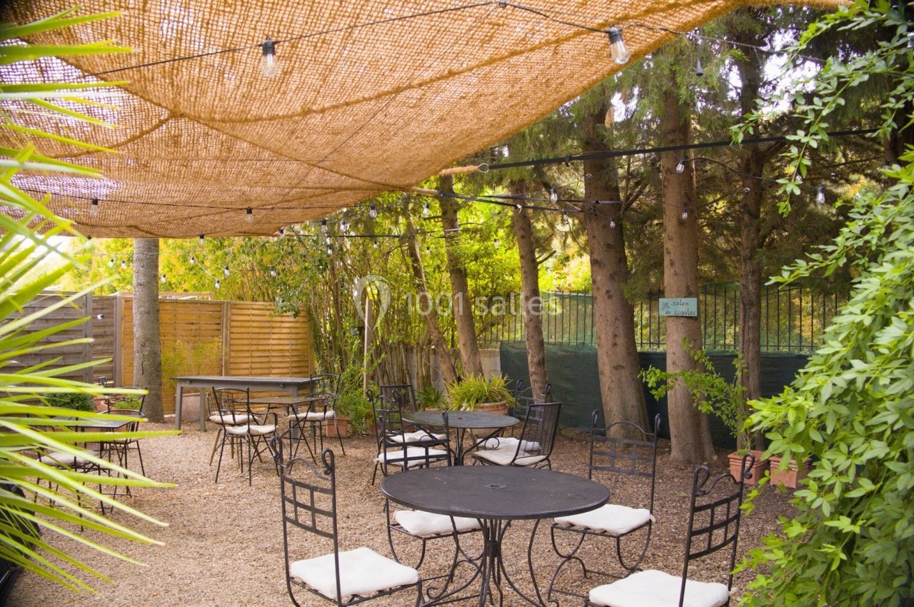 Terrasse extérieure ombragée avec tables et chaises noires, entourée de végétation et éclairée par des guirlandes lumineuses.
