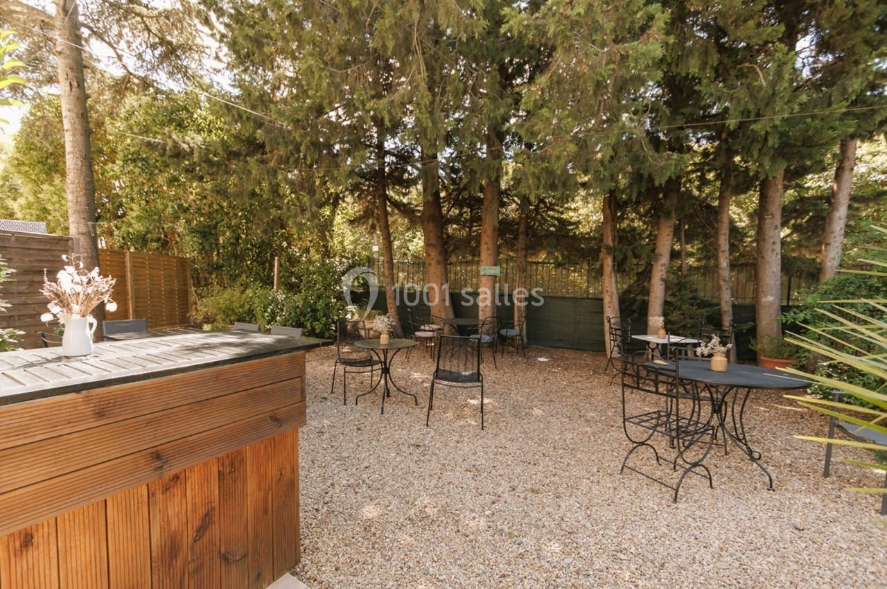 Espace extérieur avec tables et chaises sur un sol en gravier, entouré d'arbres et de verdure.