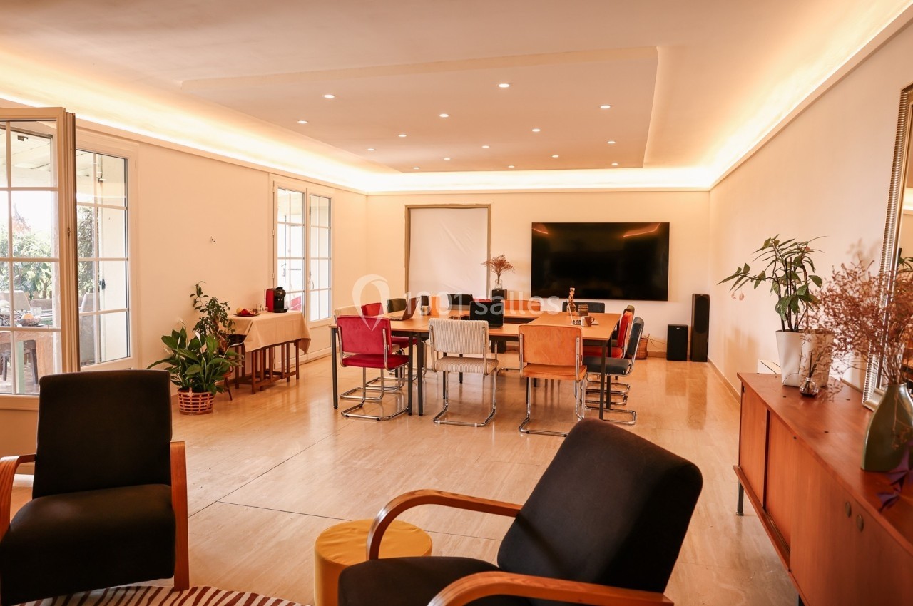 Salle à manger lumineuse avec grande table entourée de chaises colorées, écran mural et plantes décoratives.