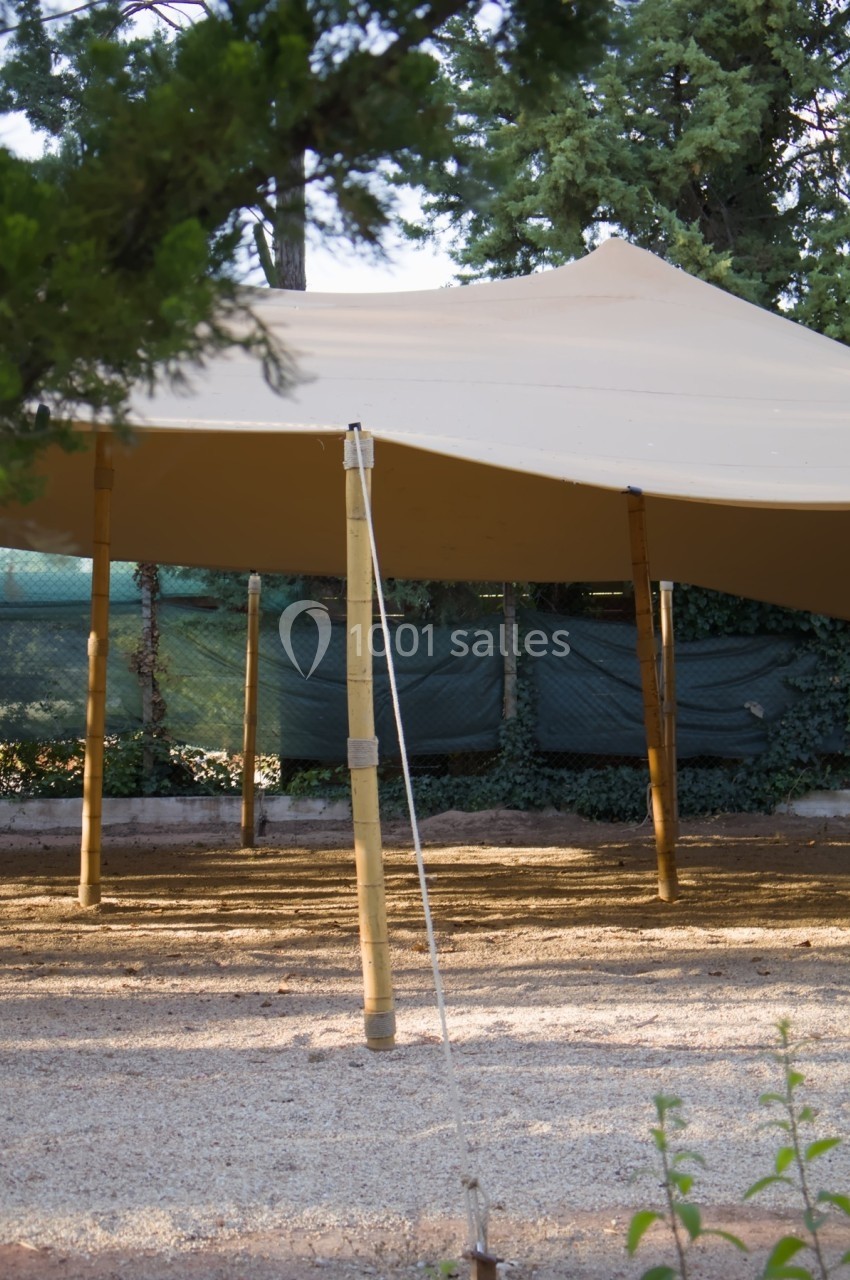 Tente beige tendue sur des poteaux en bambou, installée dans un espace extérieur entouré de végétation.