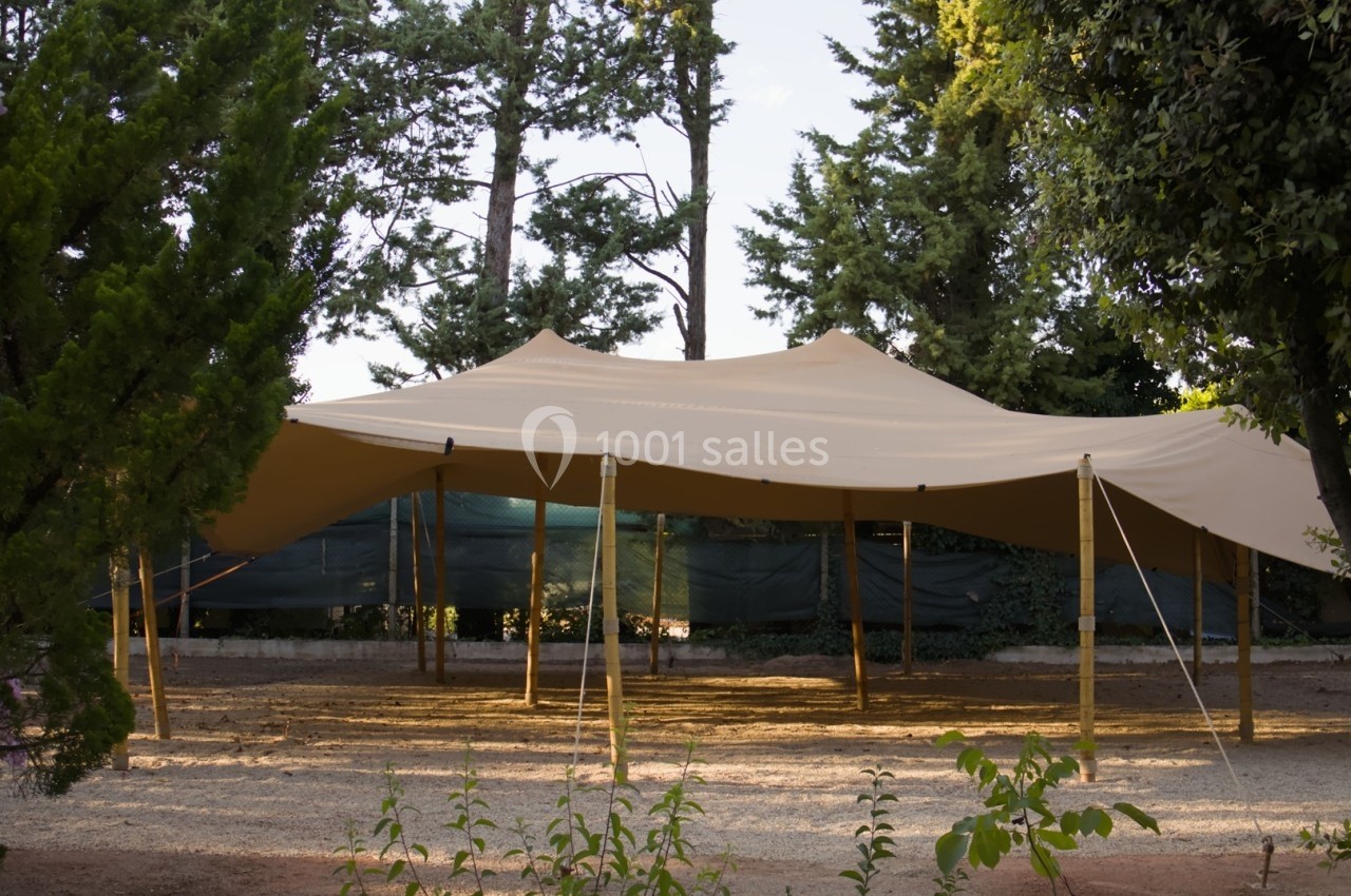 Tente beige tendue sur des poteaux en bois dans un espace extérieur entouré d'arbres et de végétation.