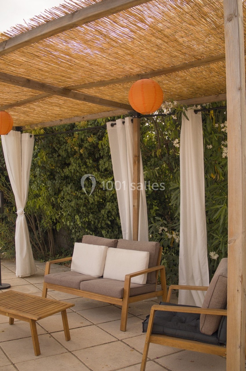 Salon de jardin en bois sous une pergola avec toit en roseaux, lanternes orange et rideaux blancs, entouré de verdure.