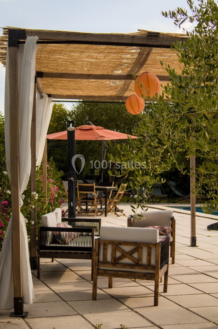 Pergola en bois avec rideaux blancs, mobilier de jardin, lanternes suspendues et parasol près d'une piscine.