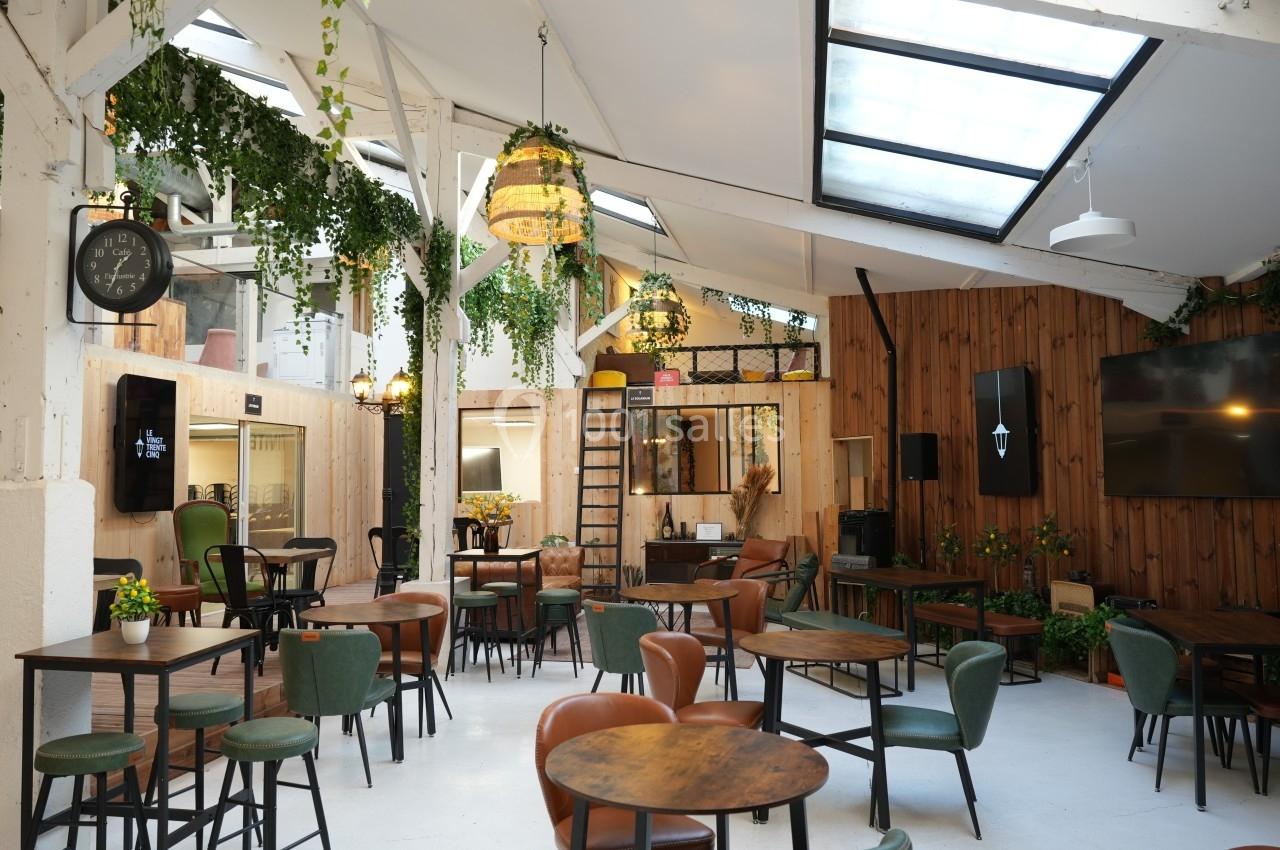 Salle lumineuse avec tables en bois, chaises variées, plantes suspendues et grandes fenêtres laissant entrer la lumière…