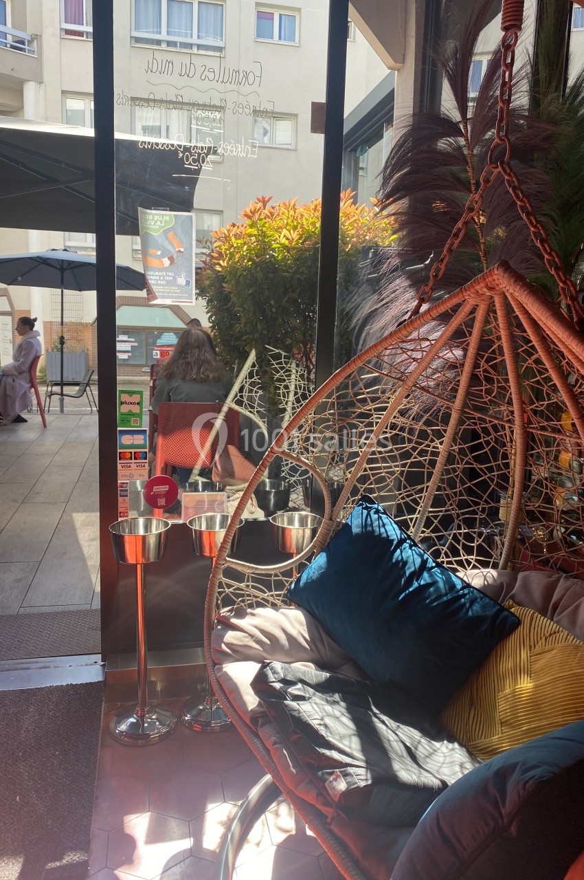 Fauteuil suspendu avec coussins colorés près d'une baie vitrée donnant sur une terrasse ensoleillée et des passants.