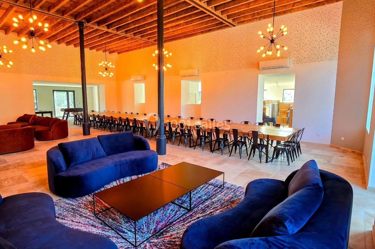 Salle spacieuse avec canapés bleus, table longue entourée de chaises, plafond en bois et luminaires suspendus.