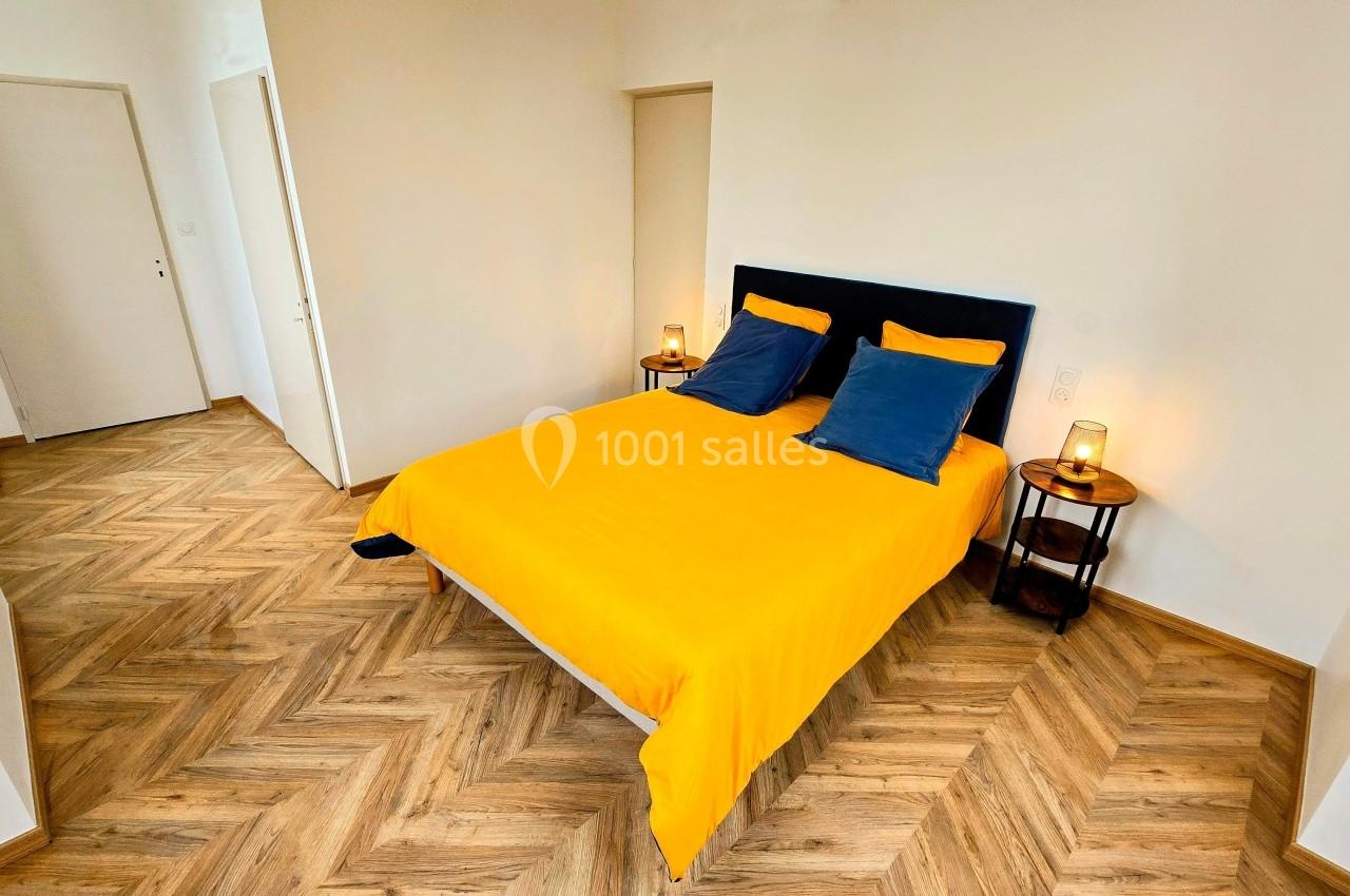 Chambre lumineuse avec lit double, housse de couette jaune, oreillers bleus et parquet en chevrons.