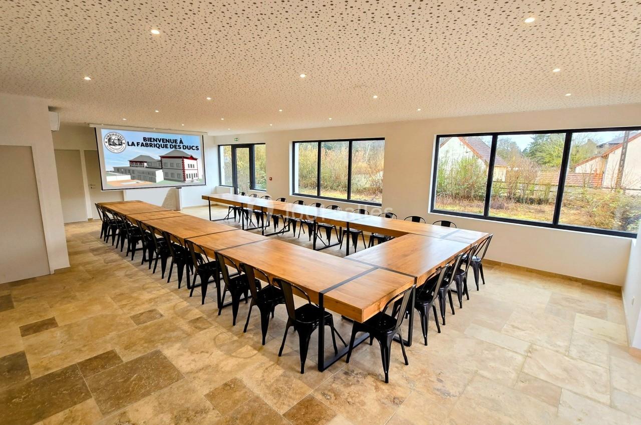 Salle de réunion lumineuse avec de grandes fenêtres, tables en bois disposées en U et chaises noires.