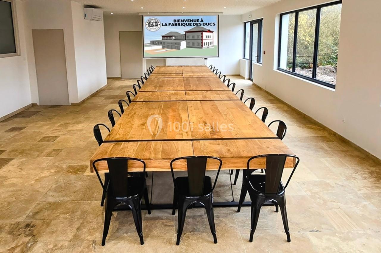 Grande salle avec une longue table en bois entourée de chaises noires, et un panneau mural indiquant ’La Fabrique des Ducs’.