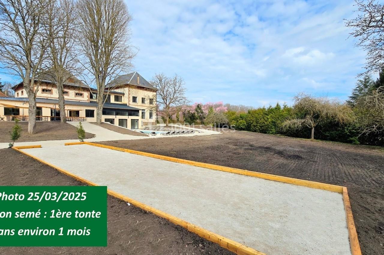 Allée en gravier bordée de bois dans un grand jardin avec pelouse ensemencée, maison en arrière-plan.