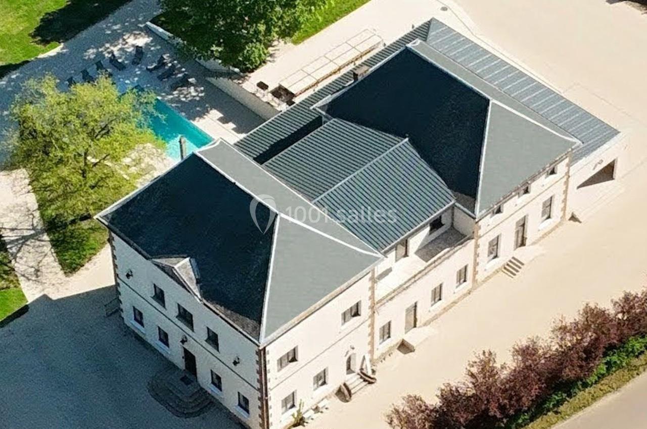Vue aérienne d'un grand bâtiment avec toit en ardoise, entouré d'arbres, d'une piscine et d'une allée pavée.