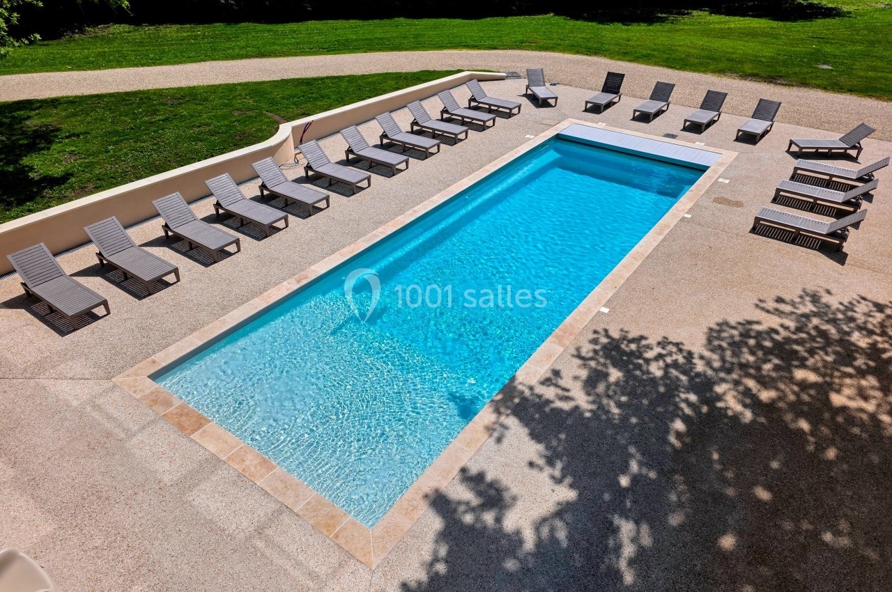 Piscine rectangulaire entourée de chaises longues sur une terrasse ensoleillée, avec pelouse et arbres en arrière-plan.