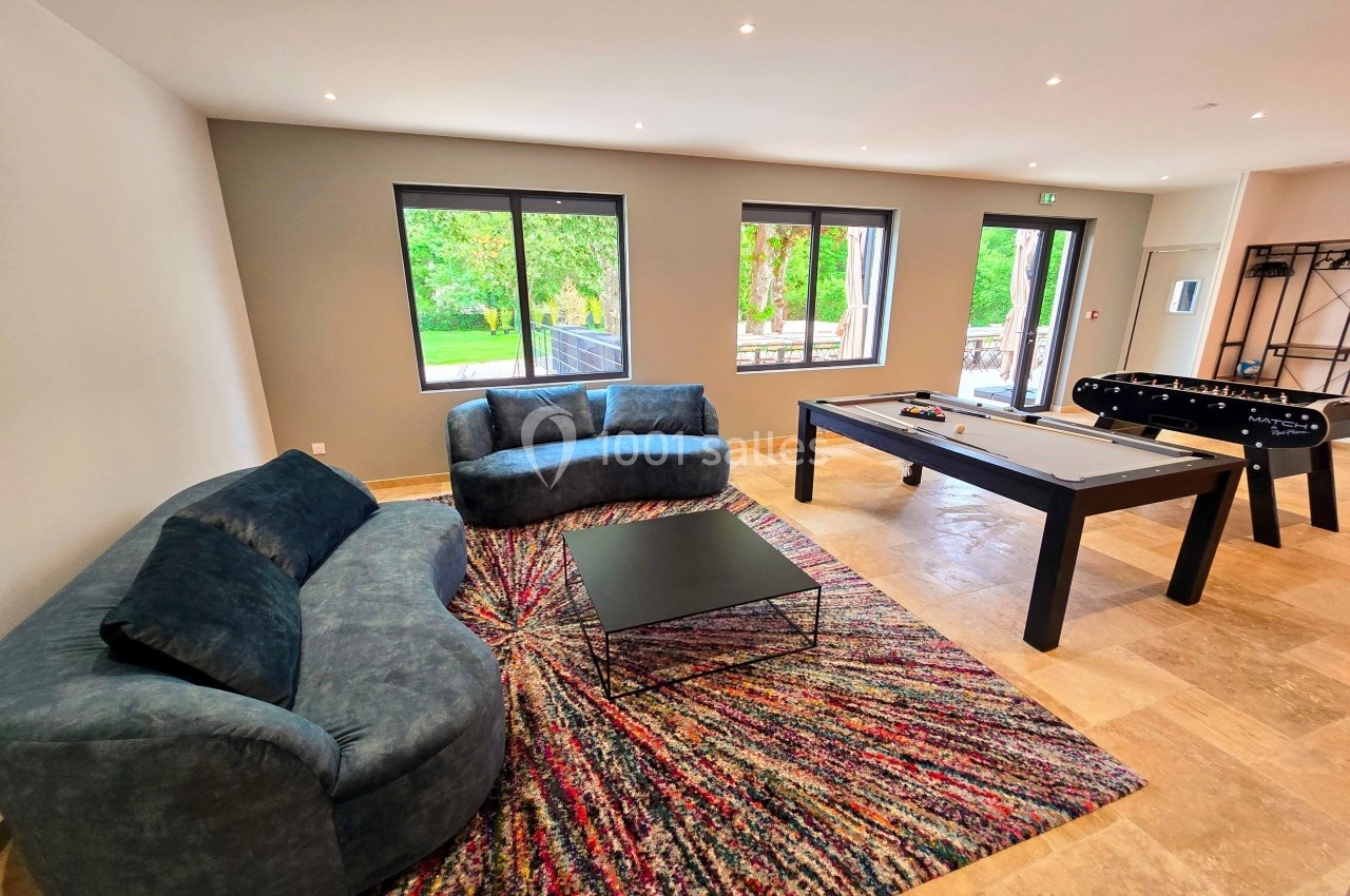 Salle de loisirs lumineuse avec canapés, tapis coloré, table de billard, baby-foot et grandes fenêtres donnant sur un jardin.