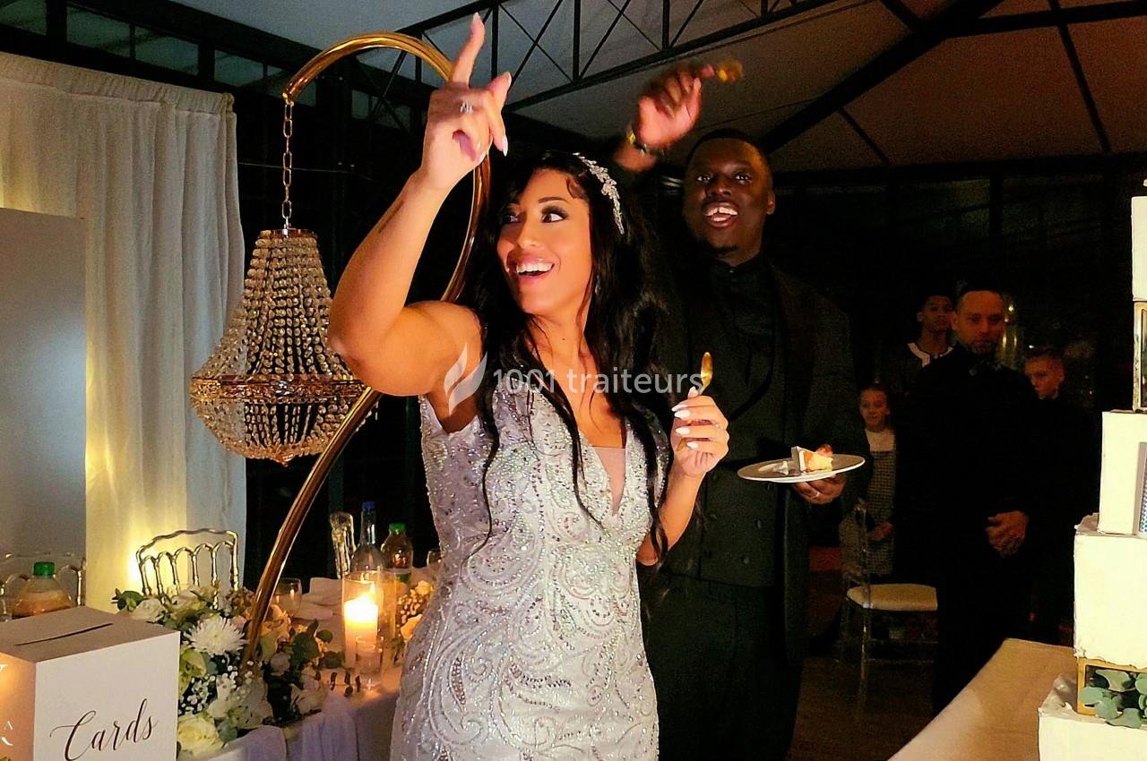 Une mariée et un invité souriants dans une salle décorée, près d'un lustre et d'un gâteau de mariage.