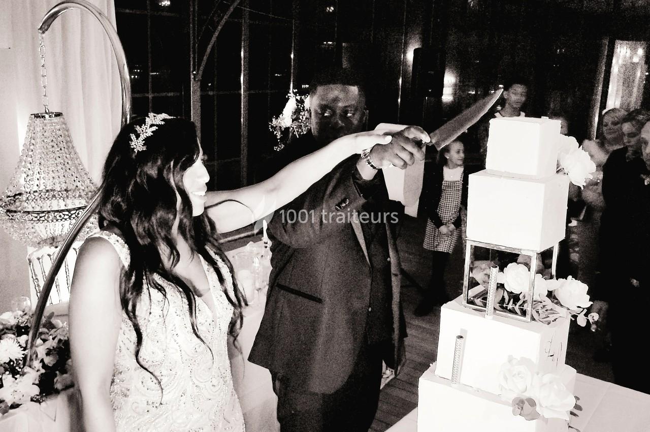 Un couple coupe un gâteau de mariage à plusieurs étages dans une salle décorée avec des fleurs et un lustre.