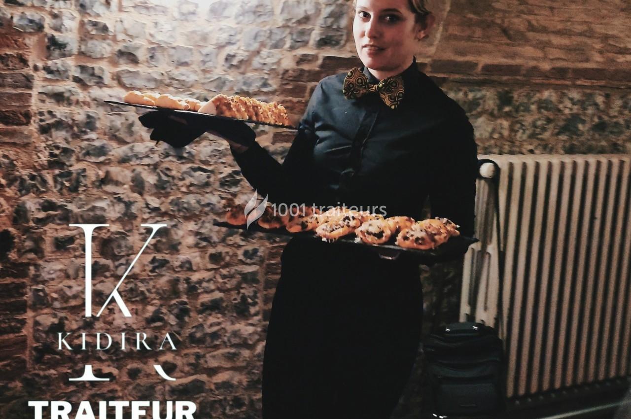 Serveuse en tenue noire portant deux plateaux de viennoiseries dans un cadre en pierre et éclairage tamisé.