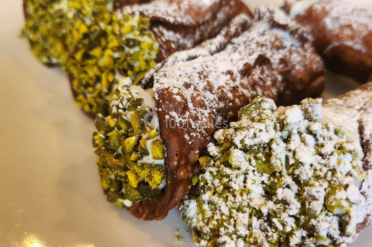 Cannoli garnis de crème, décorés de pistaches concassées et saupoudrés de sucre glace, présentés sur une assiette.