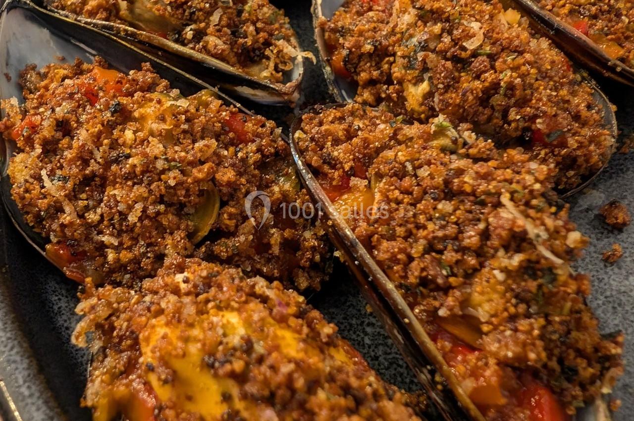 Moules gratinées garnies de chapelure dorée, herbes et morceaux de légumes, servies sur un plat en céramique.