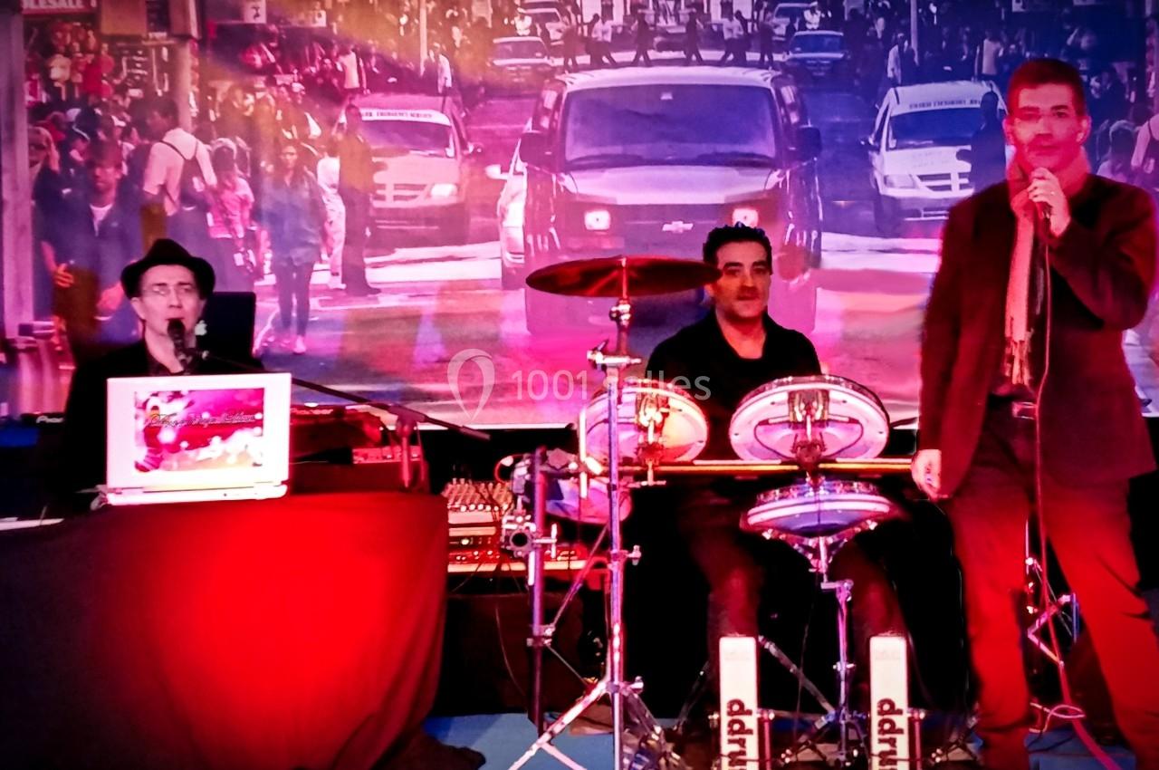 Trois musiciens sur scène, avec un chanteur, un batteur et un claviériste, devant une image de rue animée.