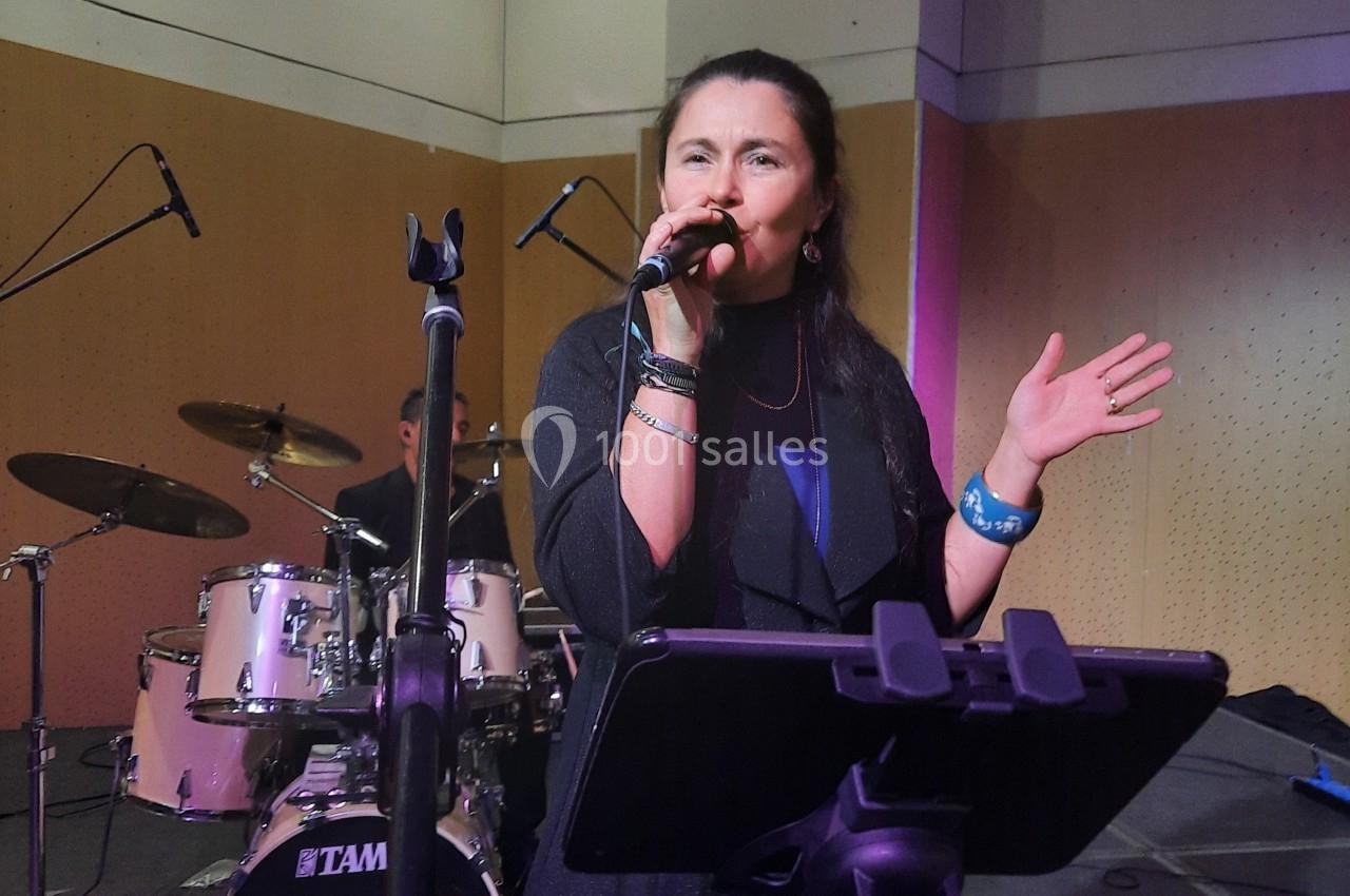 Une chanteuse sur scène interprète une chanson avec un micro, accompagnée d'un batteur en arrière-plan.