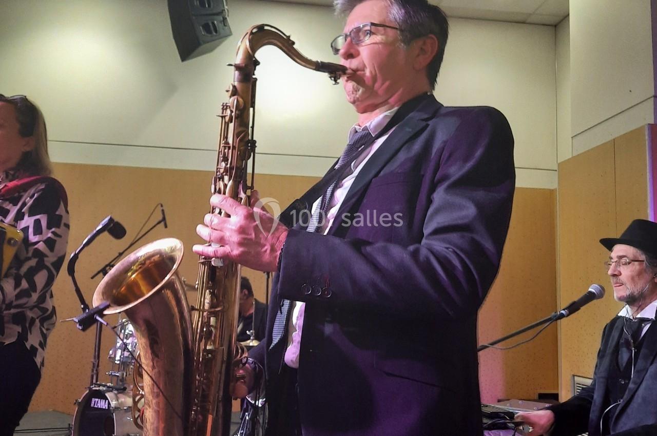 Un musicien joue du saxophone sur scène, accompagné d'un pianiste et d'une batterie en arrière-plan.