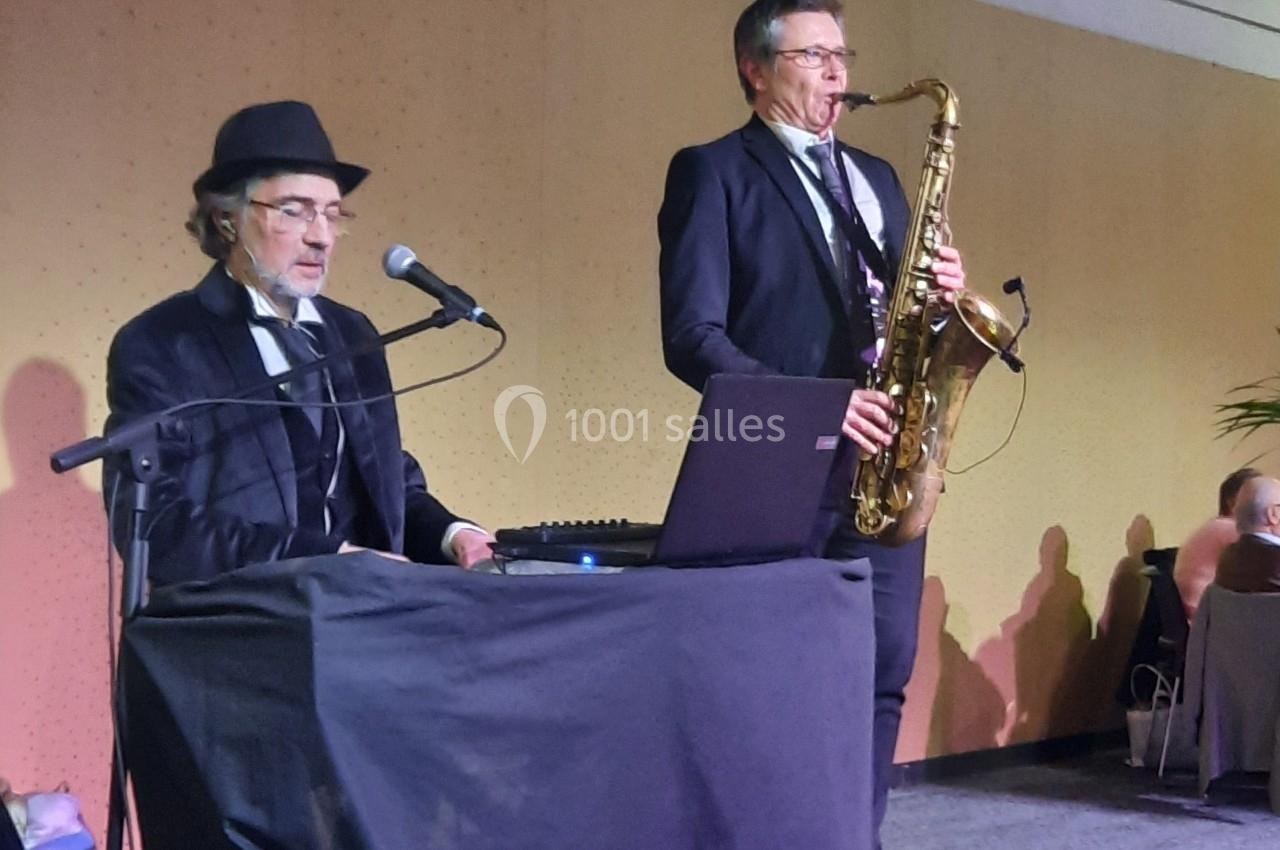 Deux musiciens sur scène, l'un joue du piano et chante, l'autre joue du saxophone, dans un cadre intérieur.