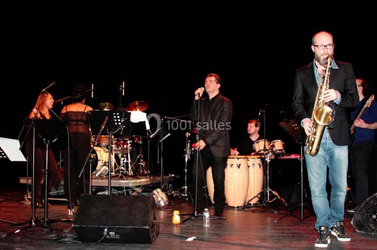 Un groupe de musiciens joue sur scène avec divers instruments, dont un saxophone, des percussions et une batterie.