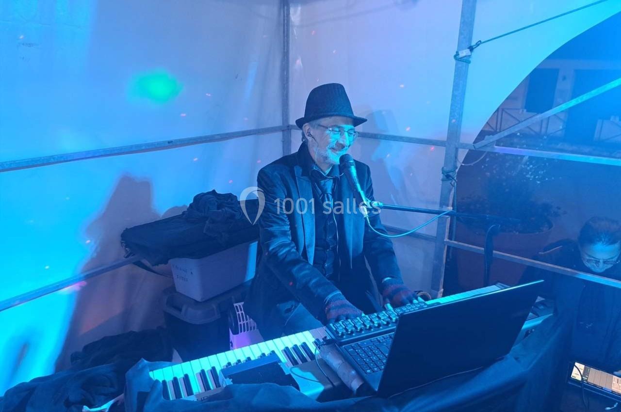 Un musicien joue du clavier et chante dans un espace éclairé de lumières bleues, portant un costume et un chapeau.