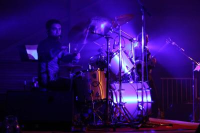 Un batteur joue sur une batterie argentée sur une scène sombre, avec un micro à proximité.