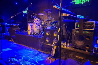 Un batteur joue sur une batterie argentée sur une scène sombre, avec un micro à proximité.