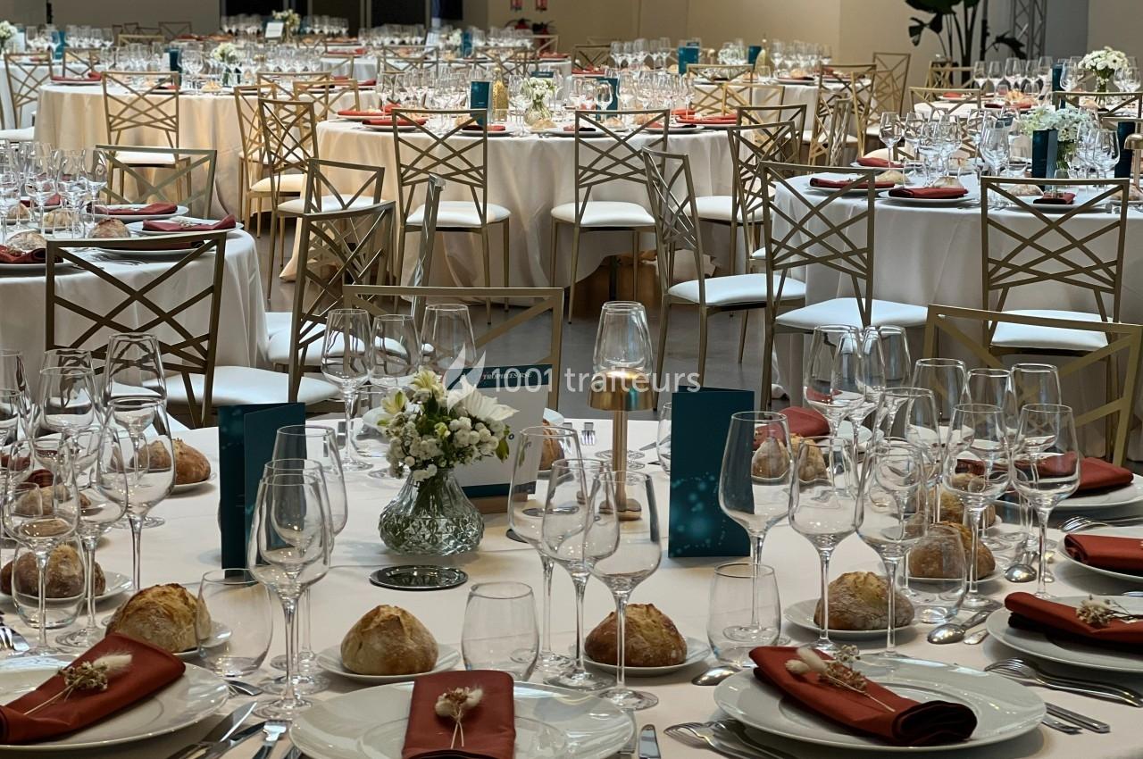 Salle de réception élégamment dressée avec des tables rondes, nappes blanches, verres, assiettes et décorations florales.