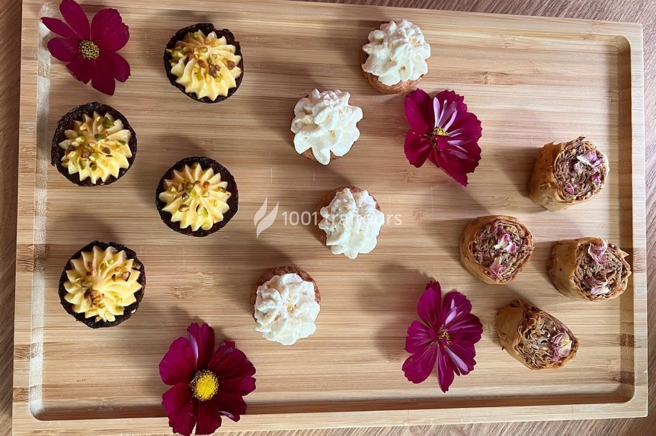 Plateau en bois avec des petits gâteaux variés décorés de crème et de fleurs comestibles, entourés de fleurs roses.