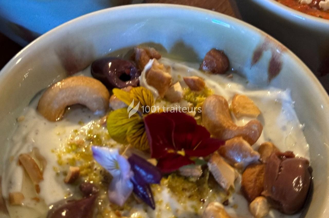 Bol de yaourt garni de noix de cajou, olives, fleurs comestibles et zestes, présenté dans une vaisselle en céramique.