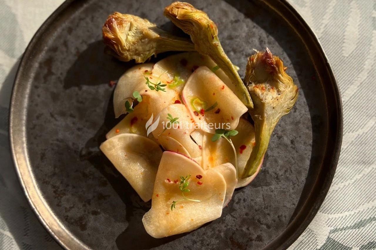 Assiette contenant des tranches fines de légumes marinés et des artichauts grillés, garnis de micro-pousses.
