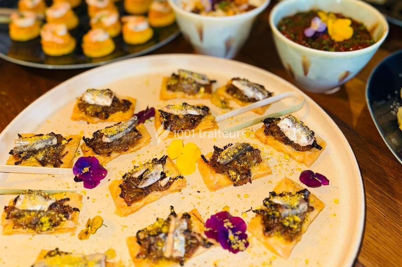 Assortiment de bouchées salées sur un plateau, décorées de fleurs comestibles, avec des plats en arrière-plan.