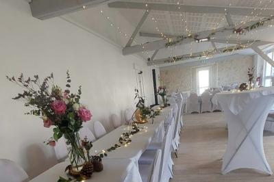 Salle décorée pour un événement avec tables blanches, chaises couvertes, guirlandes lumineuses et bouquets de fleurs.