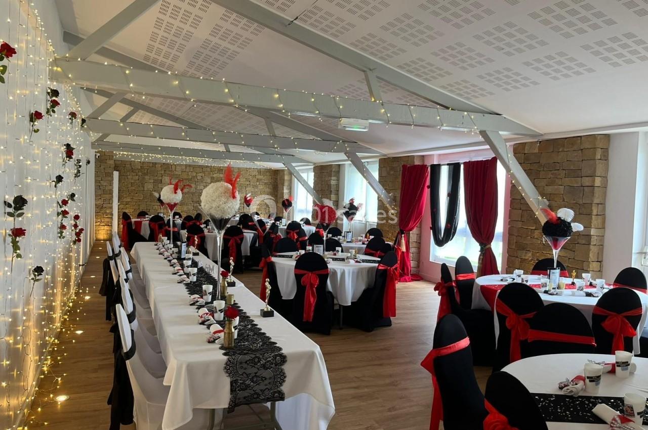 Salle de réception décorée avec des tables dressées, nappes blanches, chaises noires ornées de nœuds rouges et guirlandes…