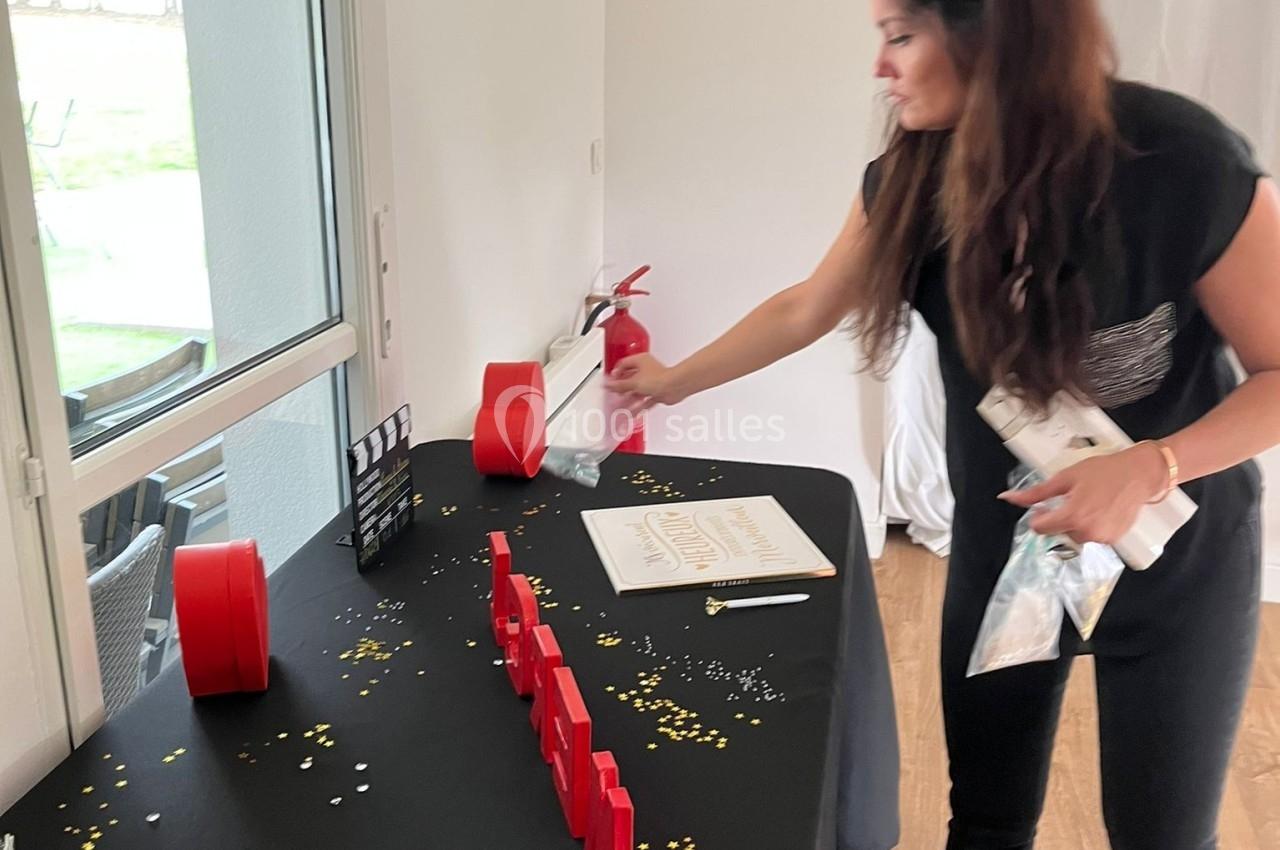 Une femme décore une table noire avec des confettis dorés et argentés, près d'une fenêtre dans une pièce lumineuse.
