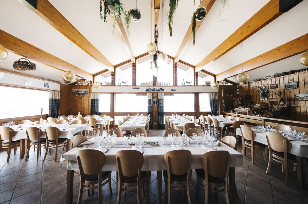 Salle de restaurant lumineuse avec de grandes baies vitrées, des tables dressées et une décoration en bois et plantes…