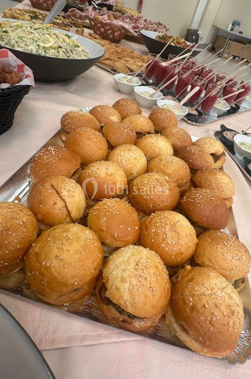 Plateau de mini-burgers au pain brioché et sésame, entouré de divers plats et desserts sur une table de buffet.