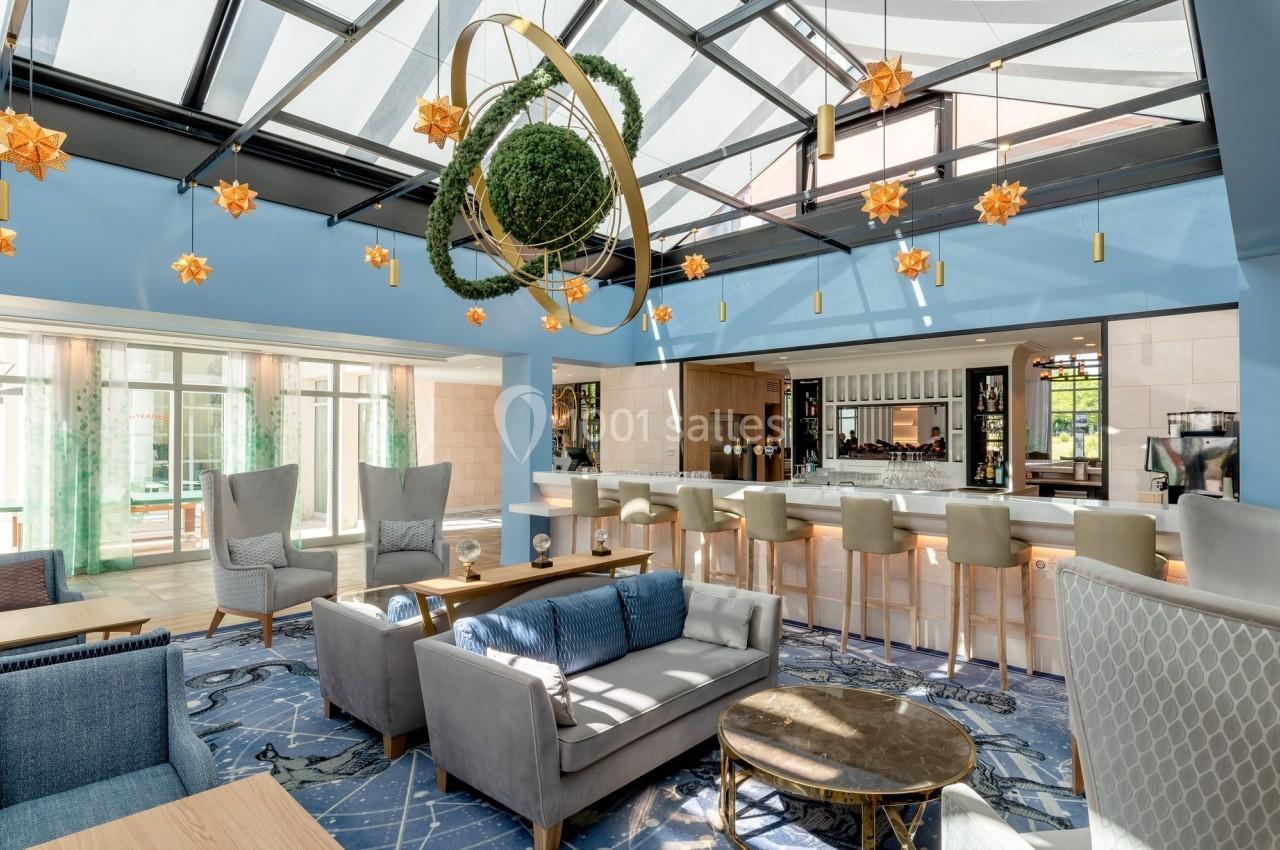 Espace lounge lumineux avec verrière, mobilier moderne, bar en arrière-plan et décoration suspendue végétale.