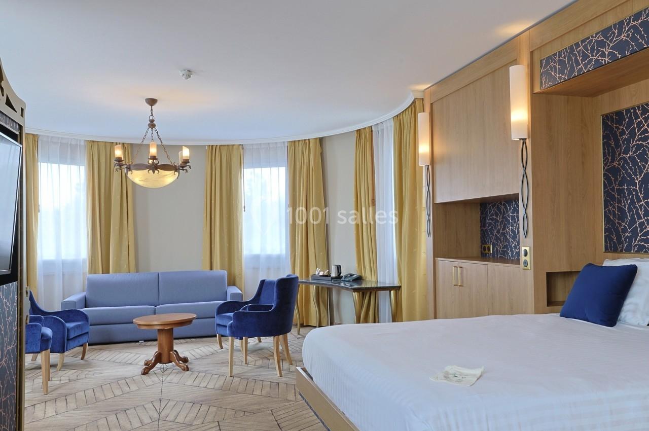 Chambre d'hôtel lumineuse avec lit double, coin salon, rideaux jaunes, mobilier en bois et fauteuils bleus.