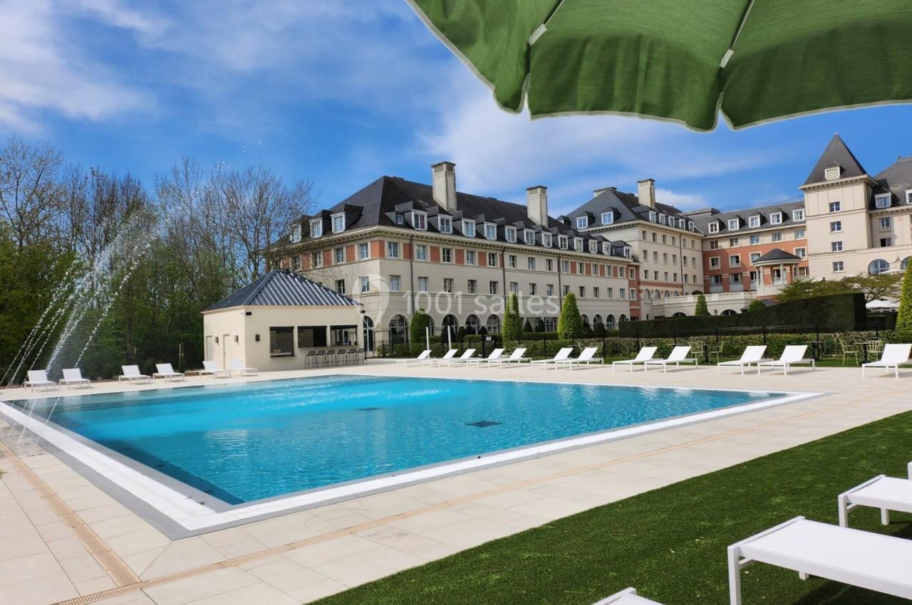 Piscine extérieure entourée de transats blancs, située devant un grand bâtiment de style classique sous un ciel bleu.
