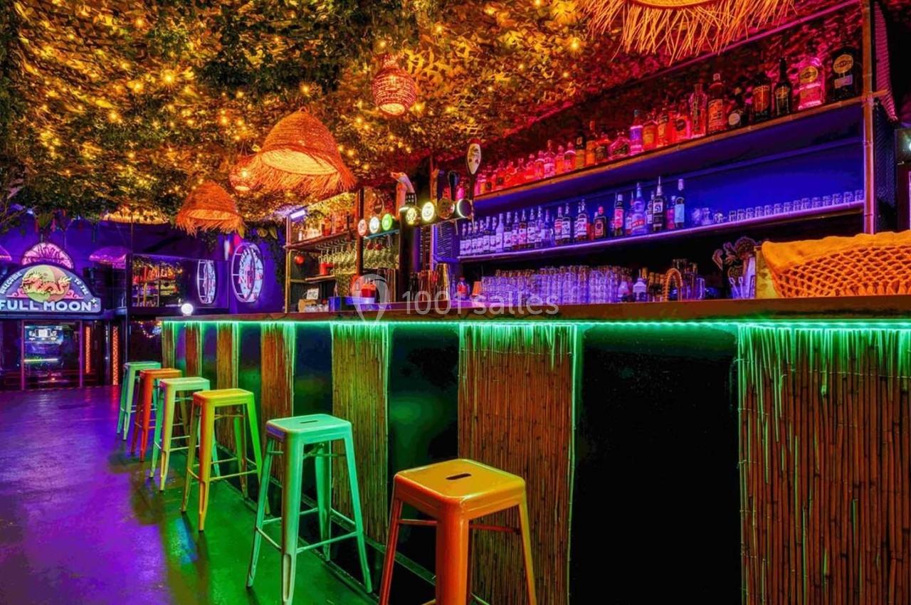 Bar coloré avec tabourets hauts, éclairage néon et plafond décoré de feuillage et lumières suspendues.