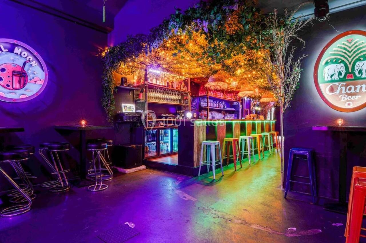 Bar coloré avec éclairage néon, tabourets hauts, comptoir illuminé et décorations végétales au plafond.