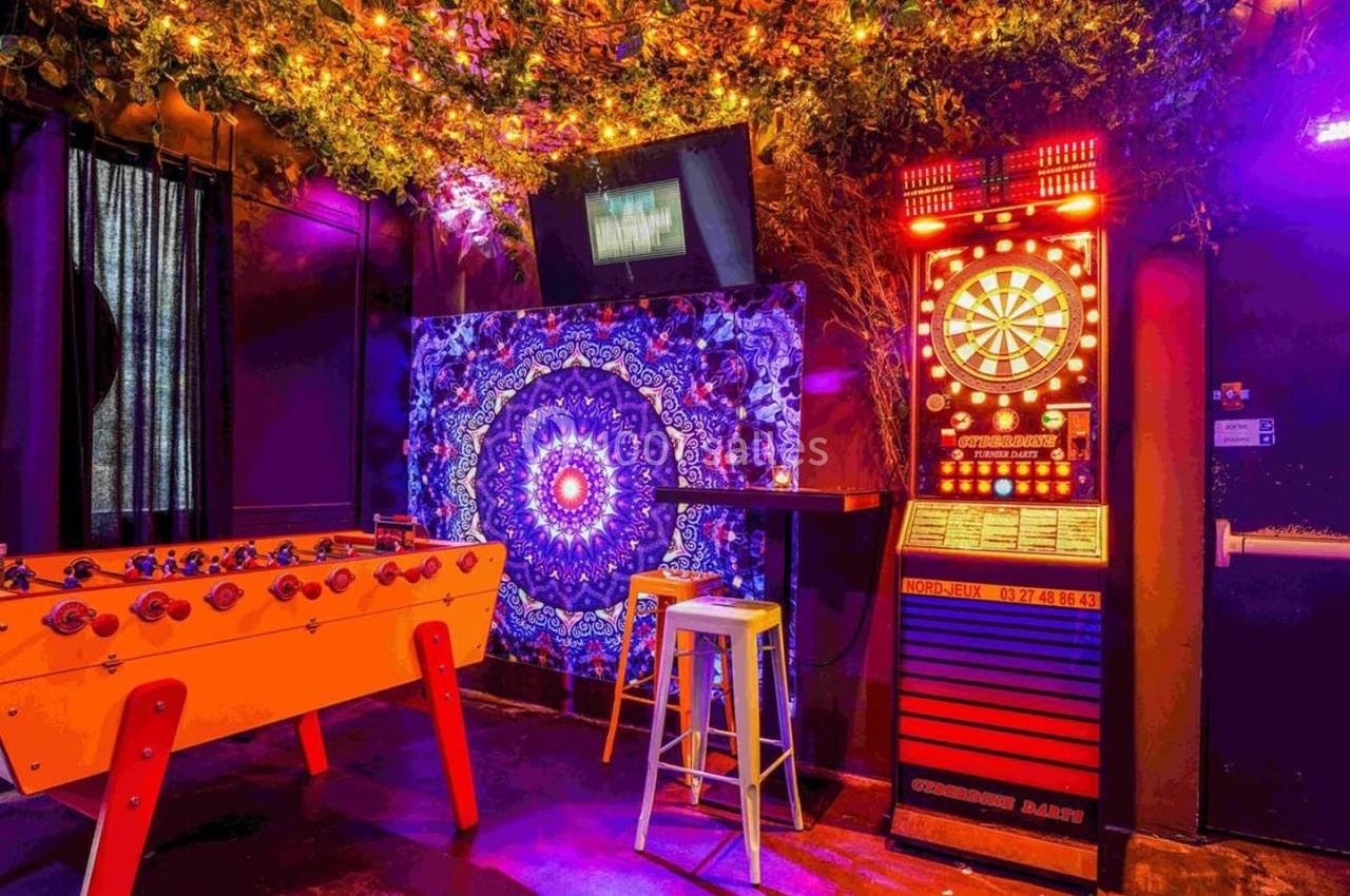Salle de loisirs avec baby-foot, jeu de fléchettes électronique, décorations lumineuses et mur orné d'un motif coloré.