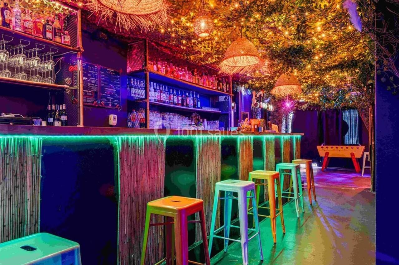 Bar coloré avec tabourets hauts, éclairage néon, décor végétal au plafond et baby-foot en arrière-plan.