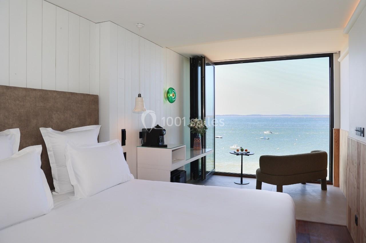 Chambre lumineuse avec lit double, fauteuil, bureau et grande baie vitrée offrant une vue sur la mer et des bateaux.