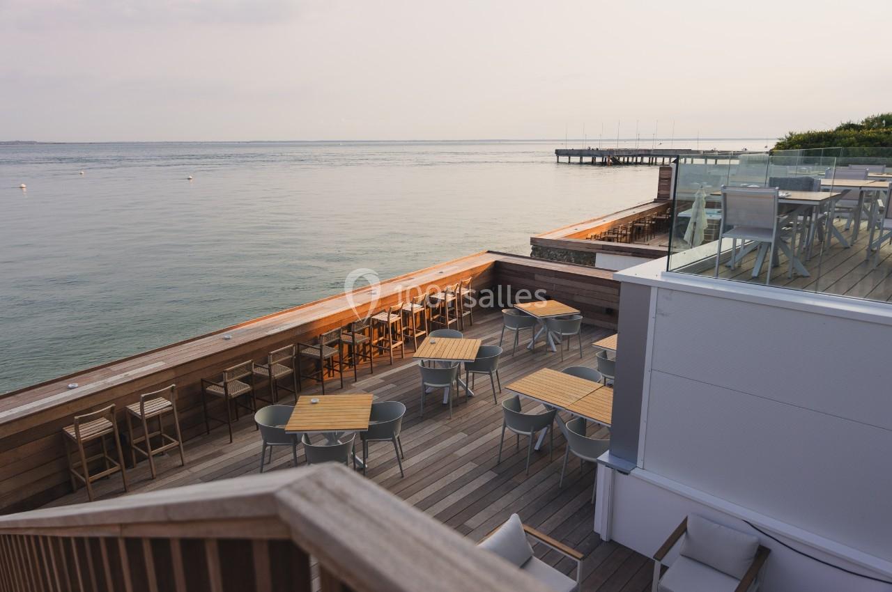 Terrasse en bois avec tables et chaises, surplombant une étendue d'eau calme sous un ciel légèrement nuageux.