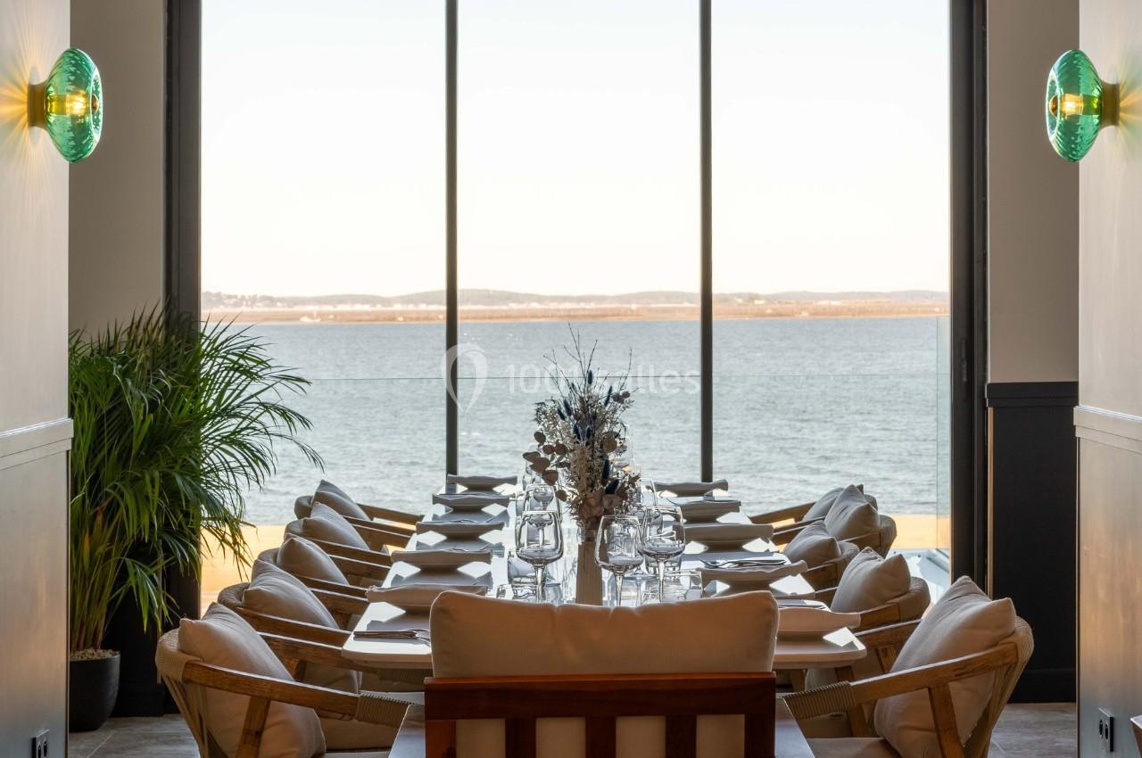 Salle à manger lumineuse avec une grande table dressée, vue sur un paysage de lac à travers de larges baies vitrées.